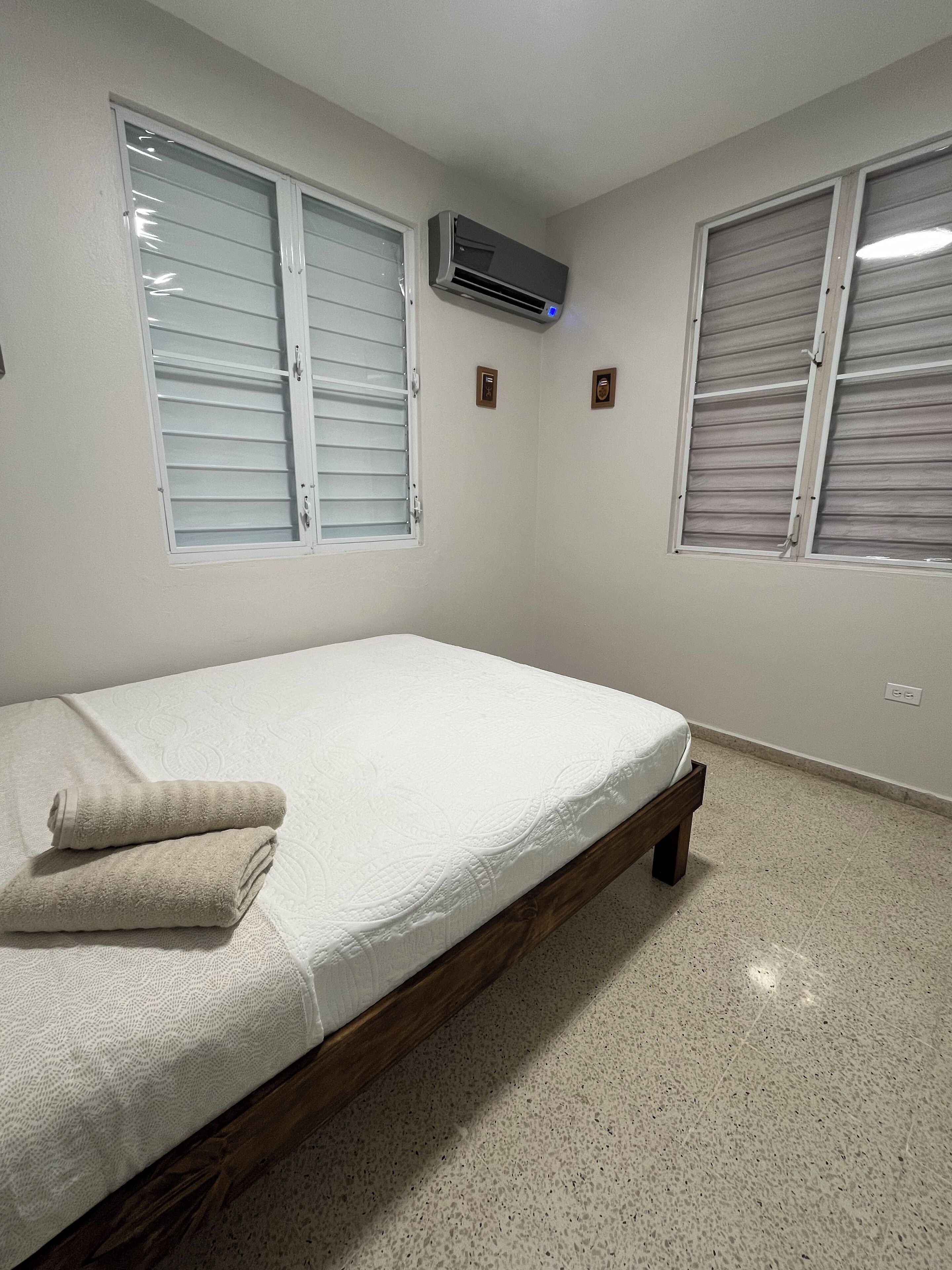 A bedroom with a bed, two windows with shutters, an air conditioning unit, and a tiled floor.