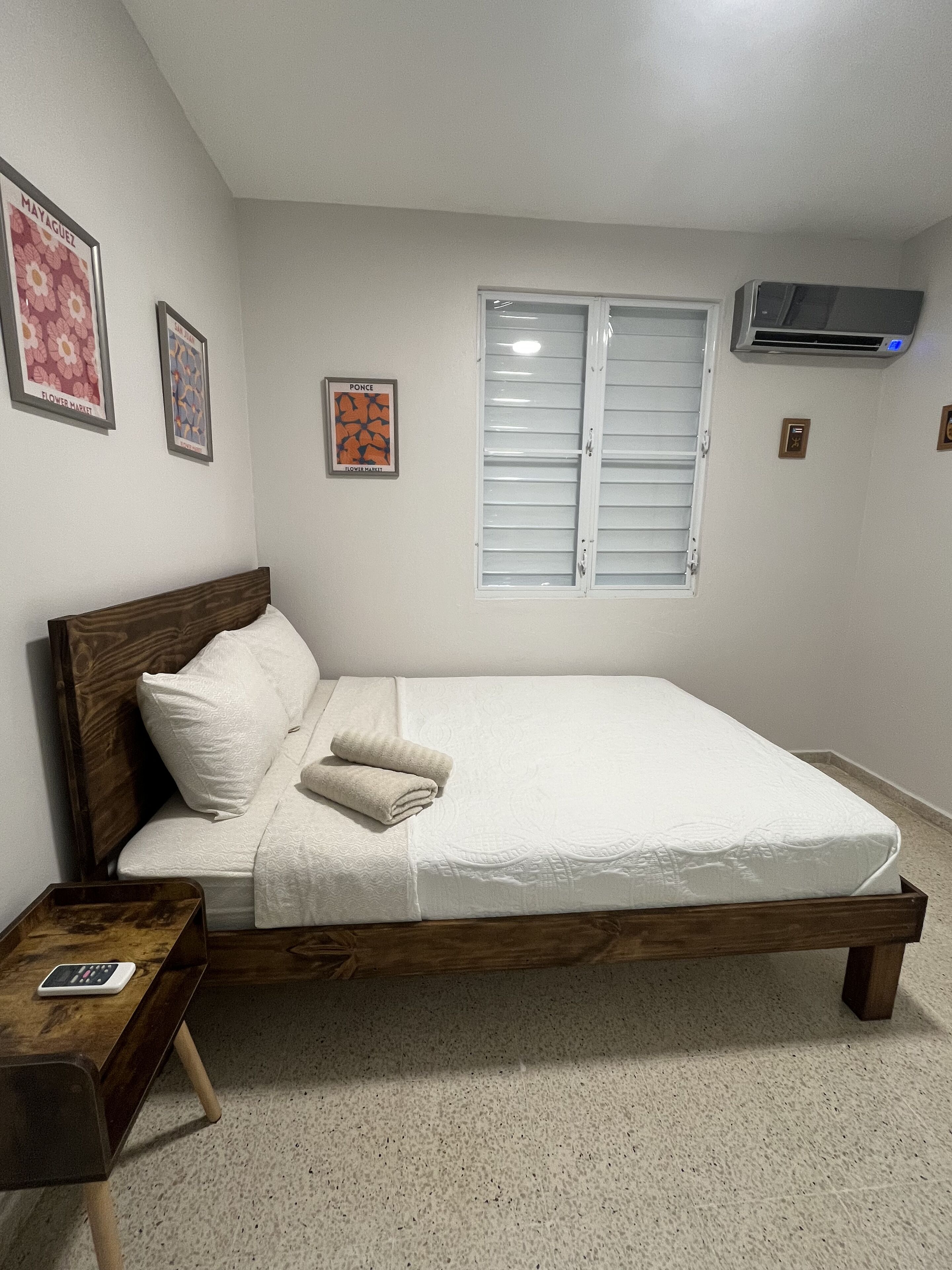 A bedroom with a wooden bed, white bedding, a bedside table, a wall-mounted air conditioner, and two framed pictures.