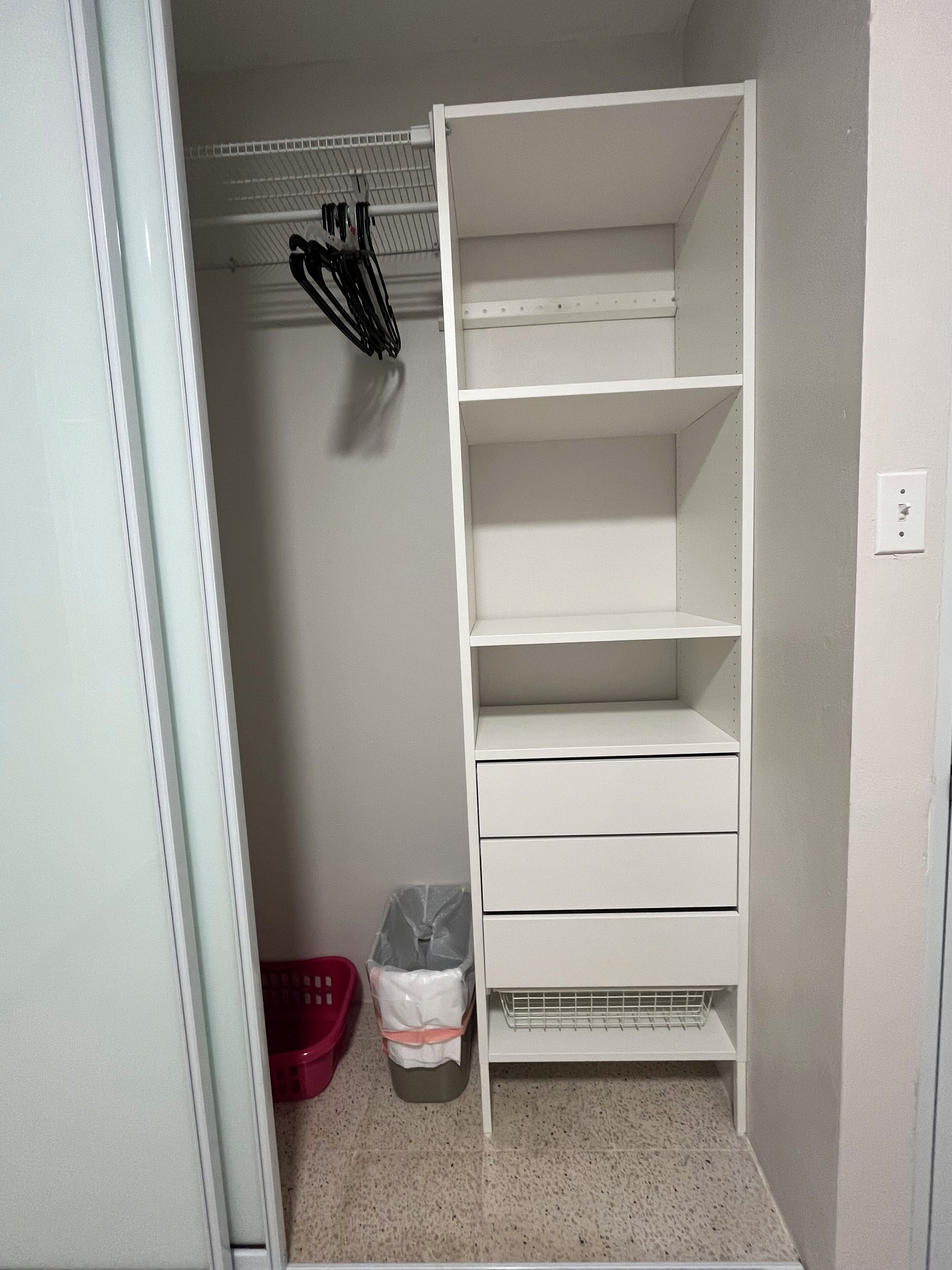 A small, white, walk-in closet with open shelves and drawers, a metal rack with hangers, and a trash bin.