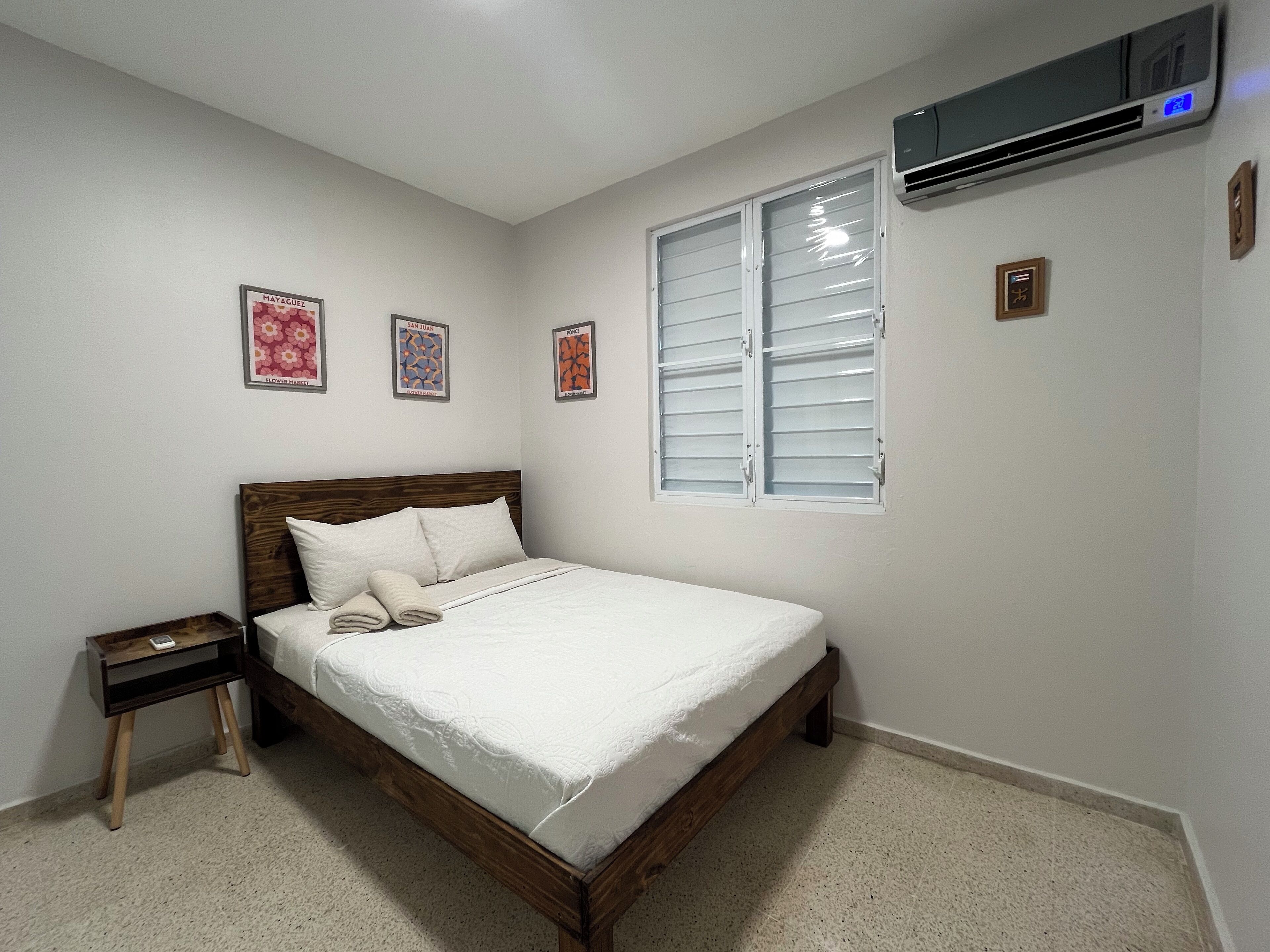 A bedroom with a wooden bed, a nightstand, a window with blinds, and three framed pictures on the wall.