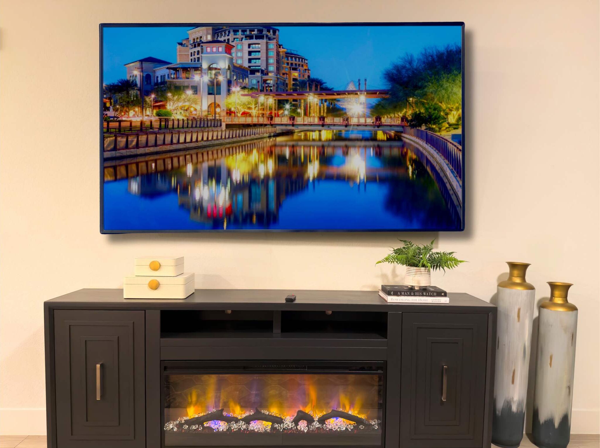 A modern living room with a flat-screen TV mounted on the wall displaying a cityscape at night. In front of the TV is a black entertainment unit with a fireplace below.