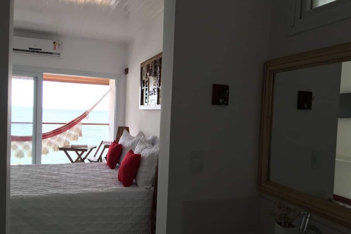 Une chambre à coucher avec un lit, une table de chevet, un miroir et une vue sur la mer par la fenêtre.