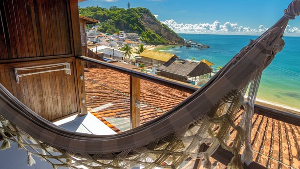 A hammock view overlooking a coastal town with terracotta roofs, a beach, and a mountainous backdrop. - POUSADA AROEIRA (Cairu)