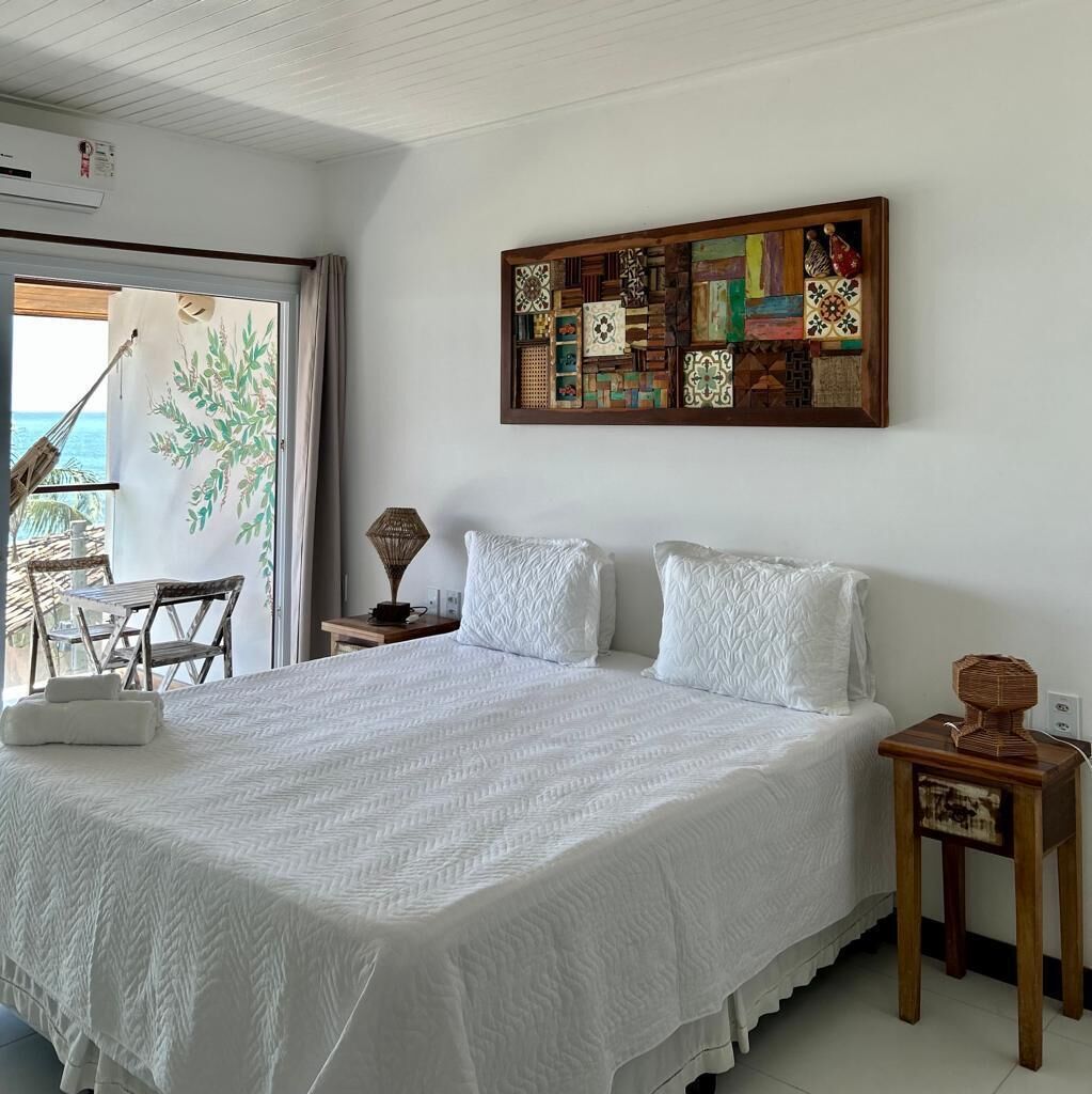 A bedroom with a large bed, two pillows, a wooden nightstand, a wall art piece, and a view of the ocean through a glass door.