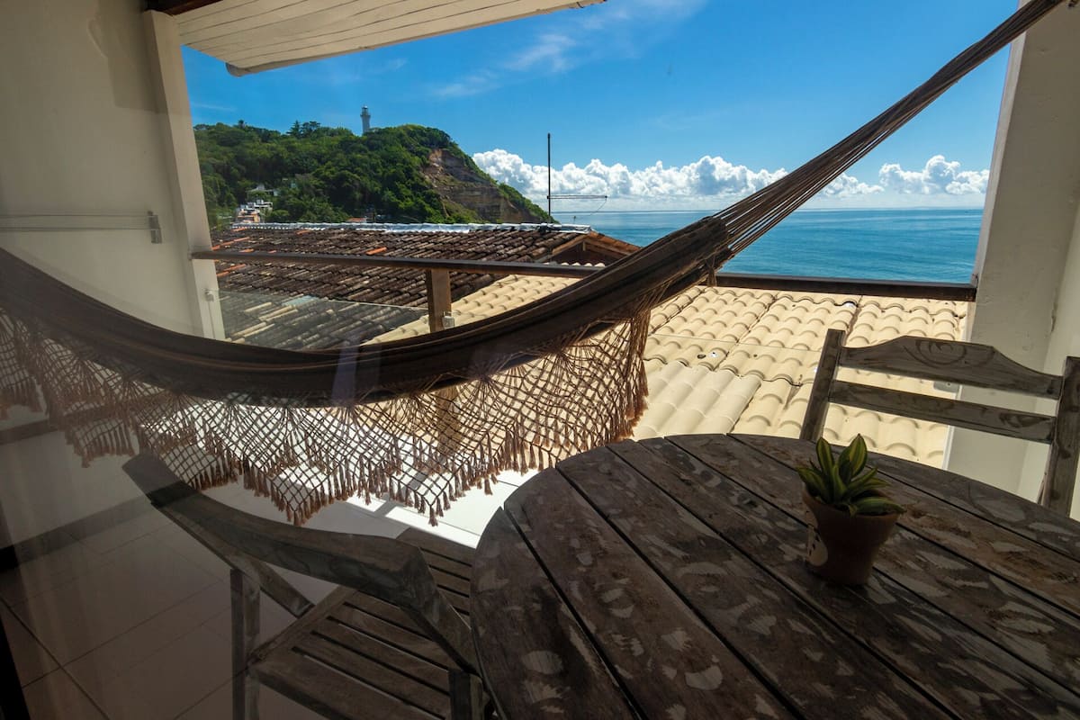 Un hamac sur un balcon donnant sur une plage, avec vue sur l’océan et un phare.