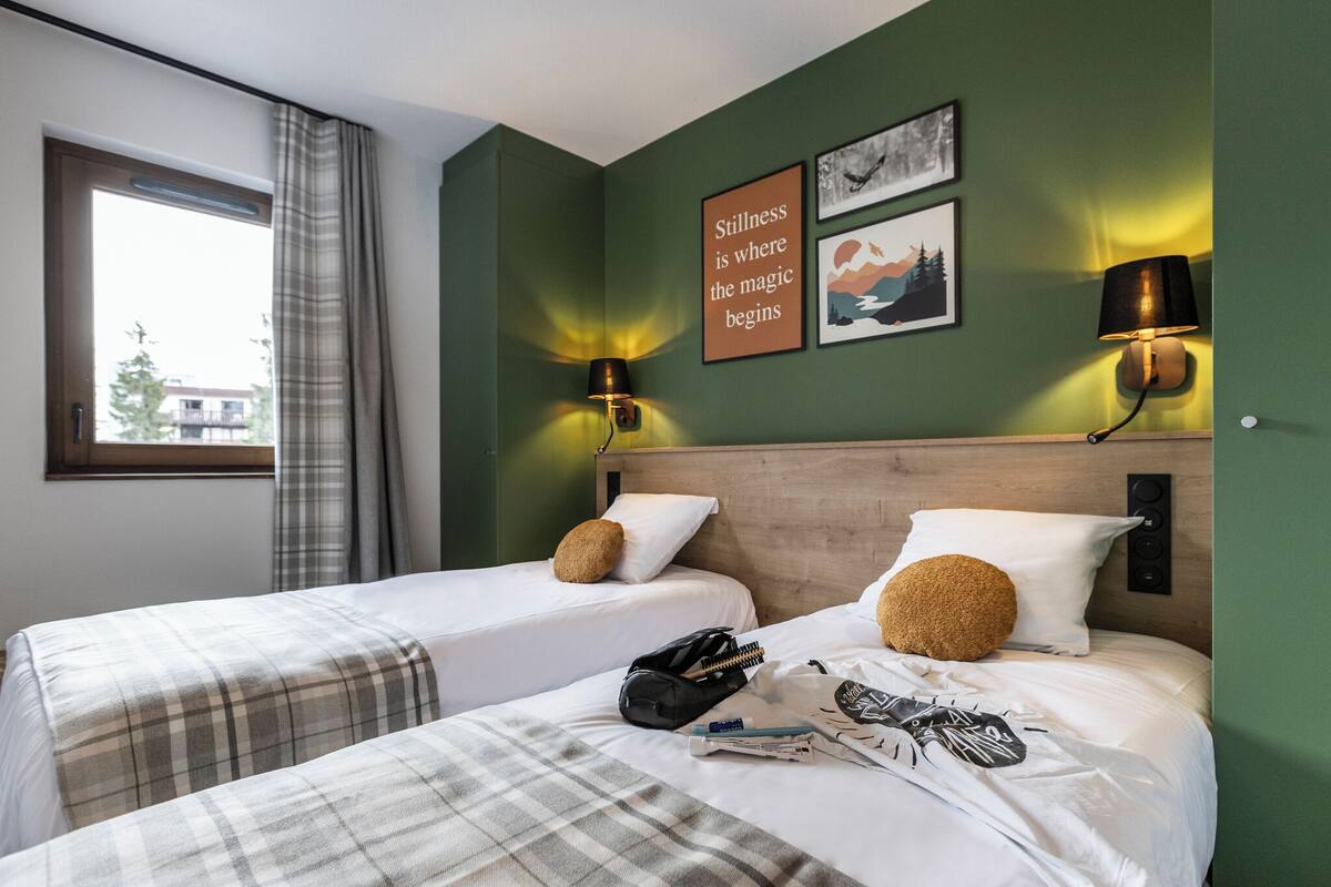 A hotel room with two beds, a window with curtains, wall art, and two wall-mounted lamps.