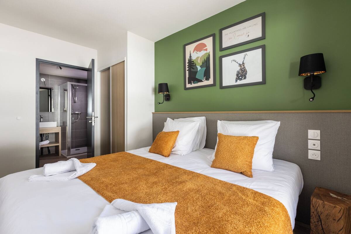 A hotel room with a bed, two wall-mounted lamps, a green wall with framed artwork, and a bathroom visible through an open door.