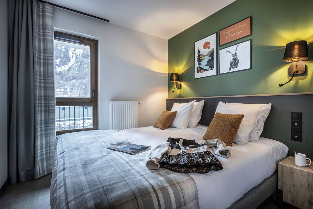 A hotel room with a bed, a window with curtains, wall-mounted lamps, and framed pictures.