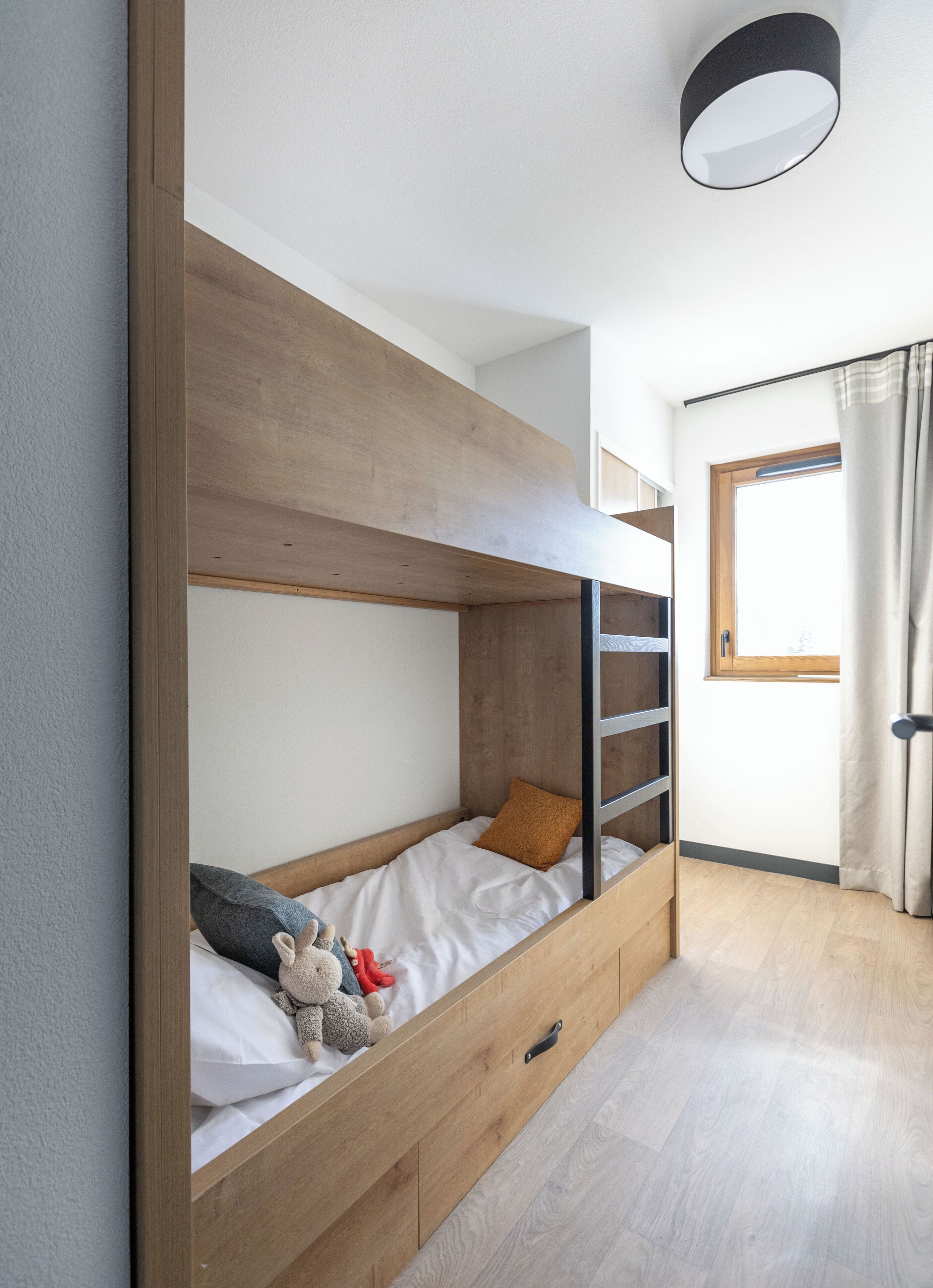 Uma cama beliche com estrutura de madeira, uma gavetinha e uma janela com cortinas.