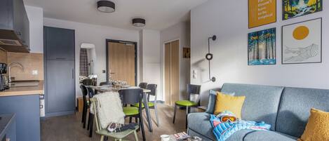 A modern living room with a sofa, coffee table, and dining area. There are framed pictures on the wall and a kitchen area in the background.