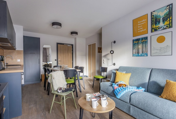 A modern living room with a sofa, coffee table, and dining area. There are framed pictures on the wall and a kitchen area in the background. - Résidence Club MMV Le Serra Neva (La Salle-les-Alpes)