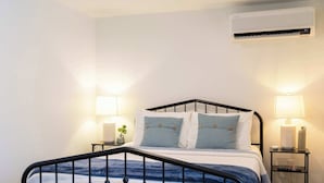 Un dormitorio con estructura metálica para la cama, una mesita de noche con lámpara y un equipo de aire acondicionado en la pared.