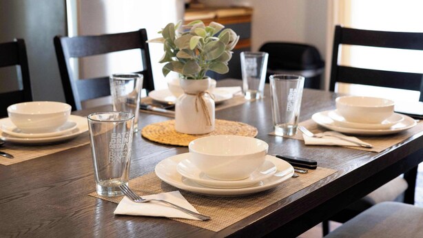 Mesa de comedor dispuesta con platos blancos, tazones y vasos transparentes, junto con una pequeña planta en maceta y un mantel tejido.