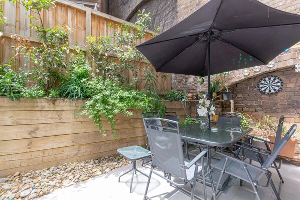 Une terrasse extérieure avec une table et des chaises, un parasol noir et un jeu de fléchettes fixé à un mur de briques.