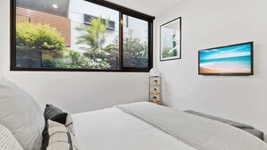Une chambre avec une grande fenêtre, un lit, une table de chevet, un téléviseur fixé au mur et un tableau encadré.