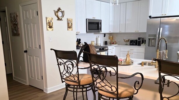Una cocina moderna con armarios blancos, electrodomésticos de acero inoxidable y una isla central. Cuatro sillas de metal con asientos acolchados rodean la isla.