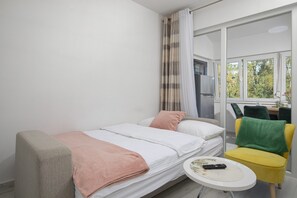 A single bed with a pink blanket, a small round table with a remote control, and a yellow chair with a green towel.