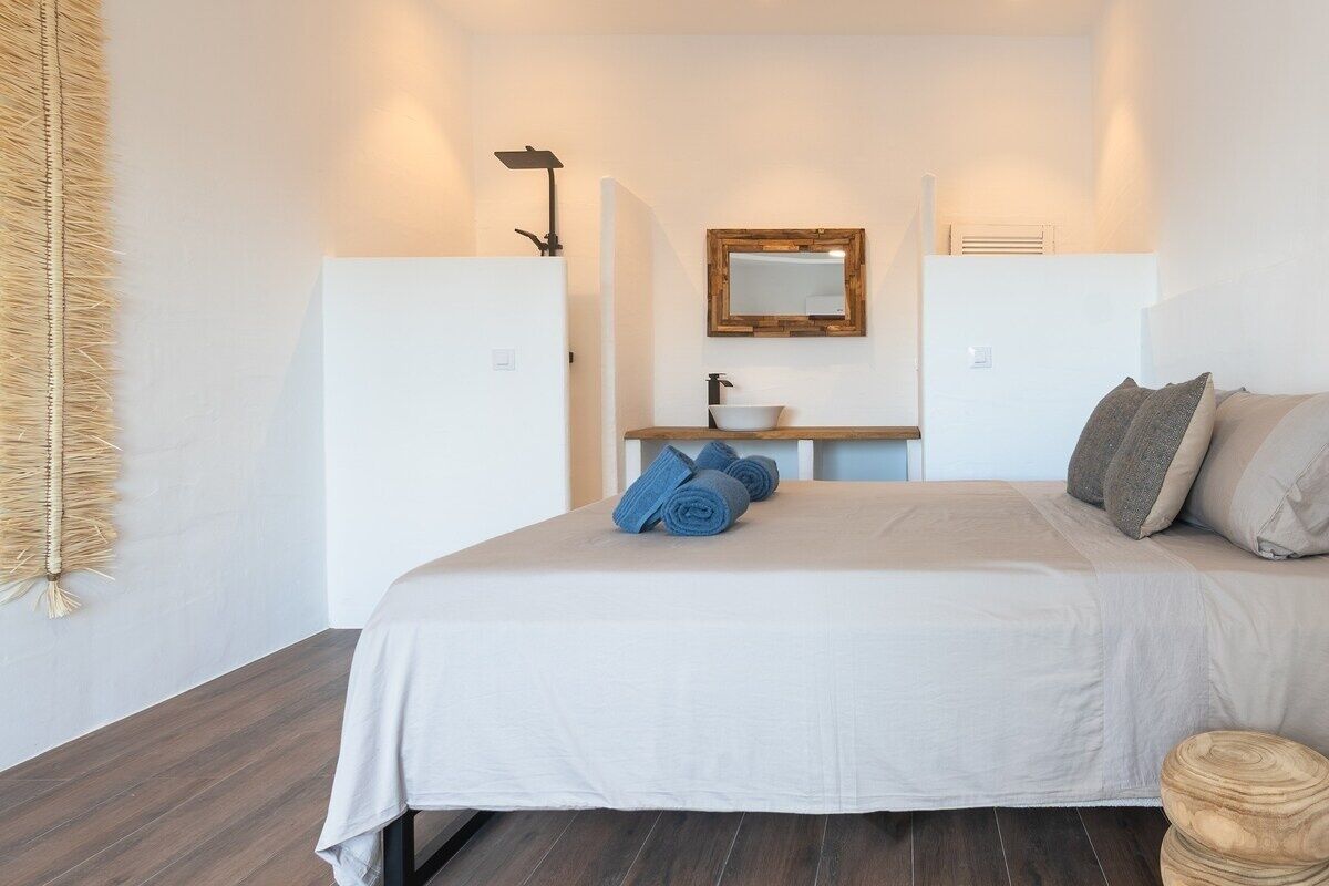 Een slaapkamer met een bed, een nachtkastje met een spiegel, een klein wastafeltje en handdoeken.