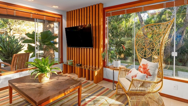 A modern living room with a TV, wicker furniture, and large windows overlooking a garden. - Bz15 Casa 200 metros da praia 4 suítes (Buzios)