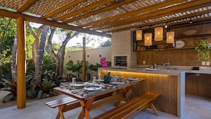 An outdoor dining area with a wooden table set for four, benches, a kitchen area with a built-in counter, and hanging pendant lights. - Bz15 Casa 200 metros da praia 4 suítes (Buzios)
