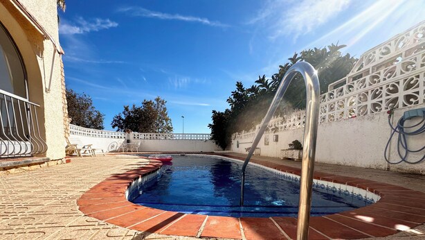 Une piscine extérieure avec un escalier en métal, entourée d’une terrasse à motifs et d’un mur décoratif blanc.