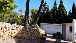 Un chemin de pierre mène à un petit bâtiment à l’extérieur blanc, entouré de palmiers et d’autres plantes vertes.