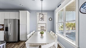 A modern kitchen with white cabinetry, a stainless steel refrigerator, a dining table with white chairs, and a window with blinds. - 4/4 - Direct Boardwalk to Beach - Neighborhood Beach Club - Beach Views (Galveston)