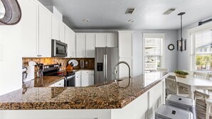 A modern kitchen with white cabinets, granite countertops, stainless steel appliances, and a window with blinds. - 4/4 - Direct Boardwalk to Beach - Neighborhood Beach Club - Beach Views (Galveston)