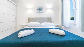 A neatly made bed with a blue bedspread and two rolled white towels, a white wall with a floral painting, and a window with curtains. - Modern apartment Marijan  (Makarska)