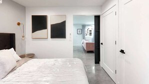 Un dormitorio moderno con una cama grande, una mesita de noche y un ventilador de techo. Hay dos cuadros enmarcados en la pared y una puerta que da a otra habitación.