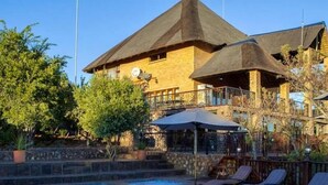A pool area with lounge chairs, a building with a unique architectural design, and a clear sky. - Lichenry Bush Lodge (Mogalakwena)