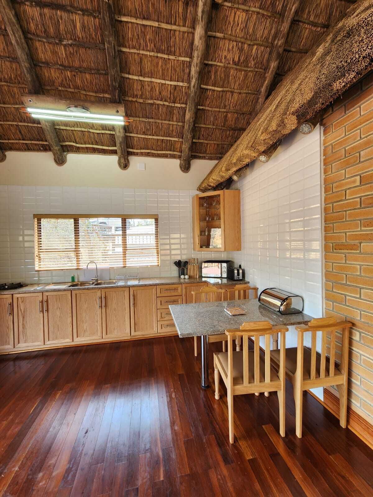 A kitchen with wooden cabinets, a granite countertop, a wooden dining table with chairs, a brick wall, and a thatched roof ceiling.