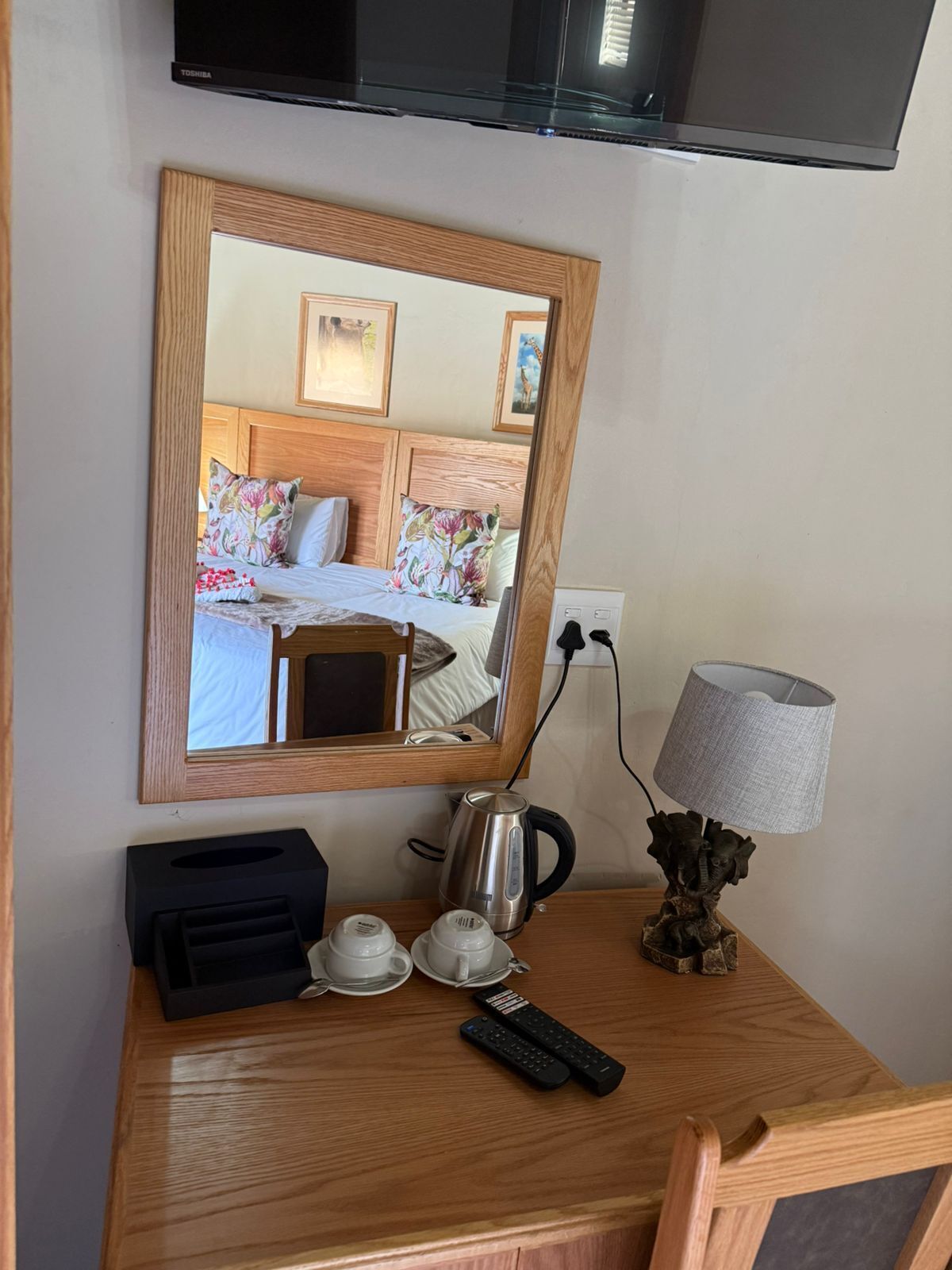 A hotel room with a wooden desk, a mirror reflecting the bed, a television, a lamp, a kettle, and two cups.