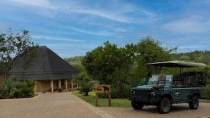 A thatched-roof building with a paved driveway leading to a green safari vehicle. - Lichenry Bush Lodge (Mogalakwena)
