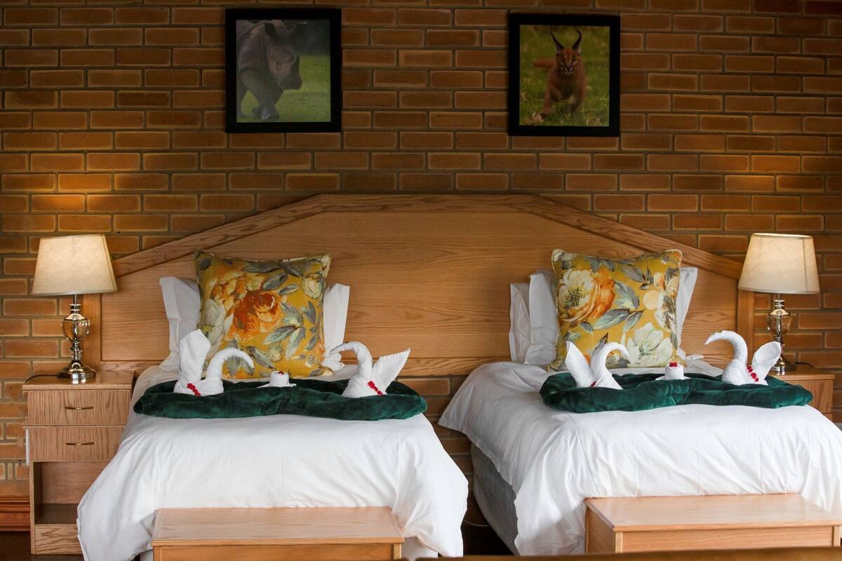 A hotel room with two beds, each with a floral pillow and a swan decoration. There are two nightstands with lamps, and the walls are brick with two framed pictures.