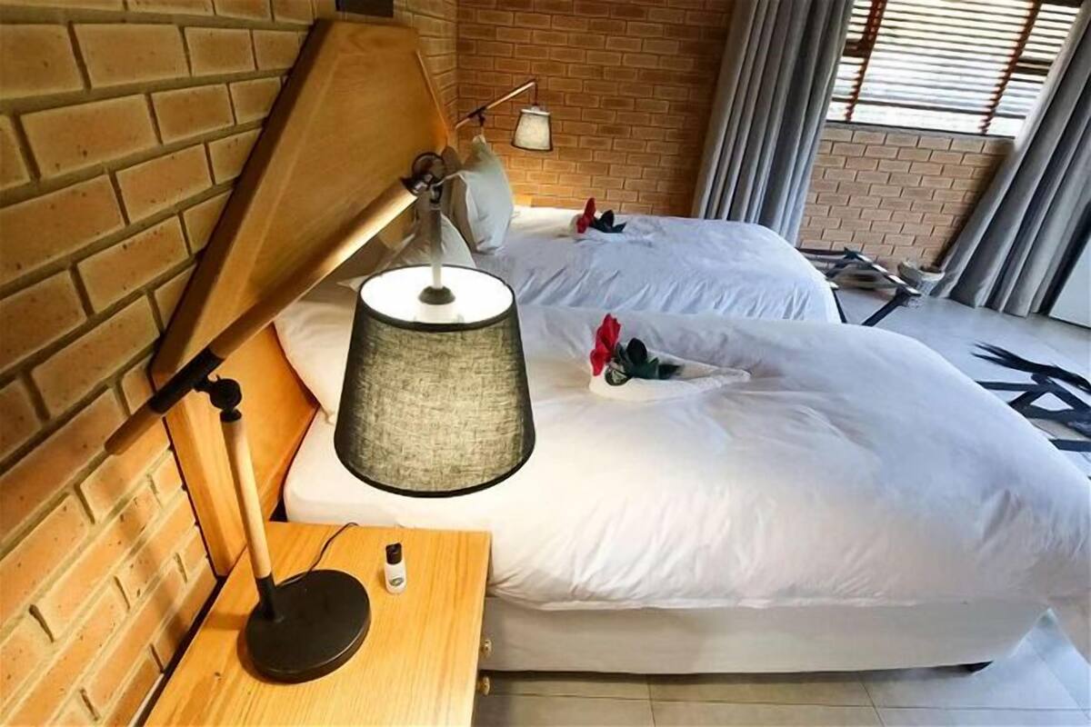 A hotel room with a brick wall, a wooden headboard, a bedside lamp, a bed with white bedding, and a small table with a bottle.