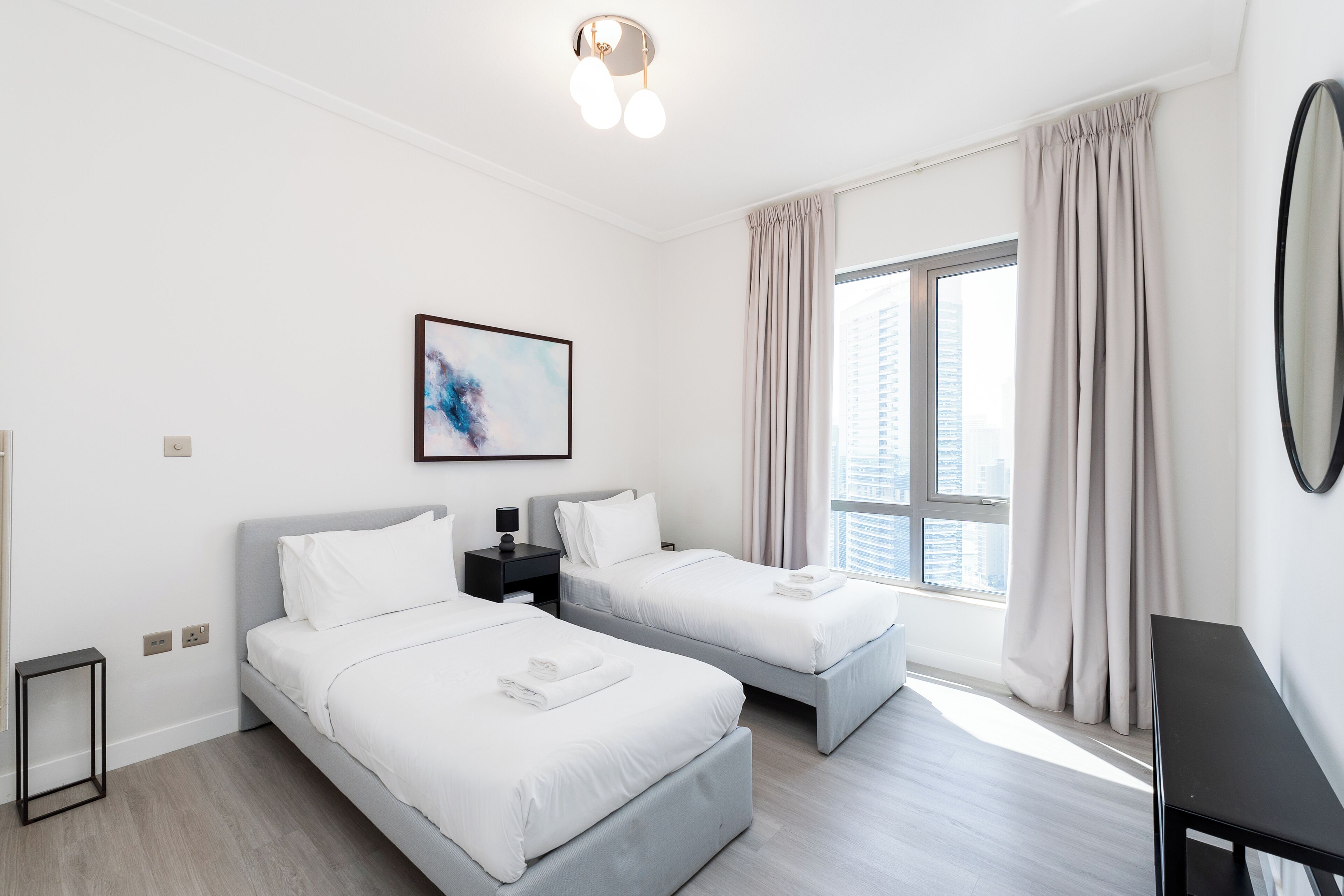 A hotel room with two beds, a large window with curtains, a painting on the wall, and a small table.
