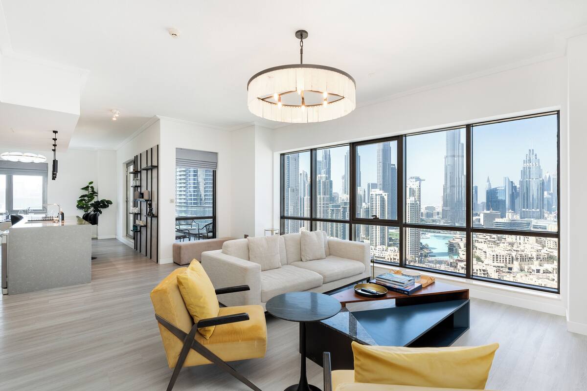 Una sala de estar moderna con sofá, sillones y una mesa de centro, ventanas amplias con vistas a la ciudad y una zona de cocina.