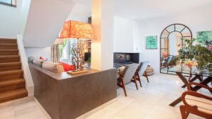 A modern living room with a grey sofa, a glass coffee table, and a TV. There is a staircase on the left and a dining area with wooden chairs. - Genteel Home Casa Aloha Golf (Marbella)