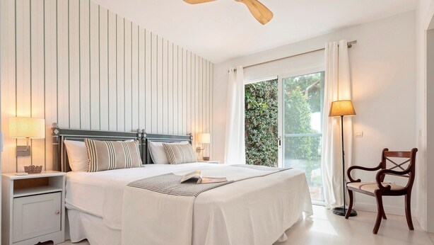 A bedroom with a bed, nightstands, a chair, a ceiling fan, and a window with curtains. - Genteel Home Casa Aloha Golf (Marbella)