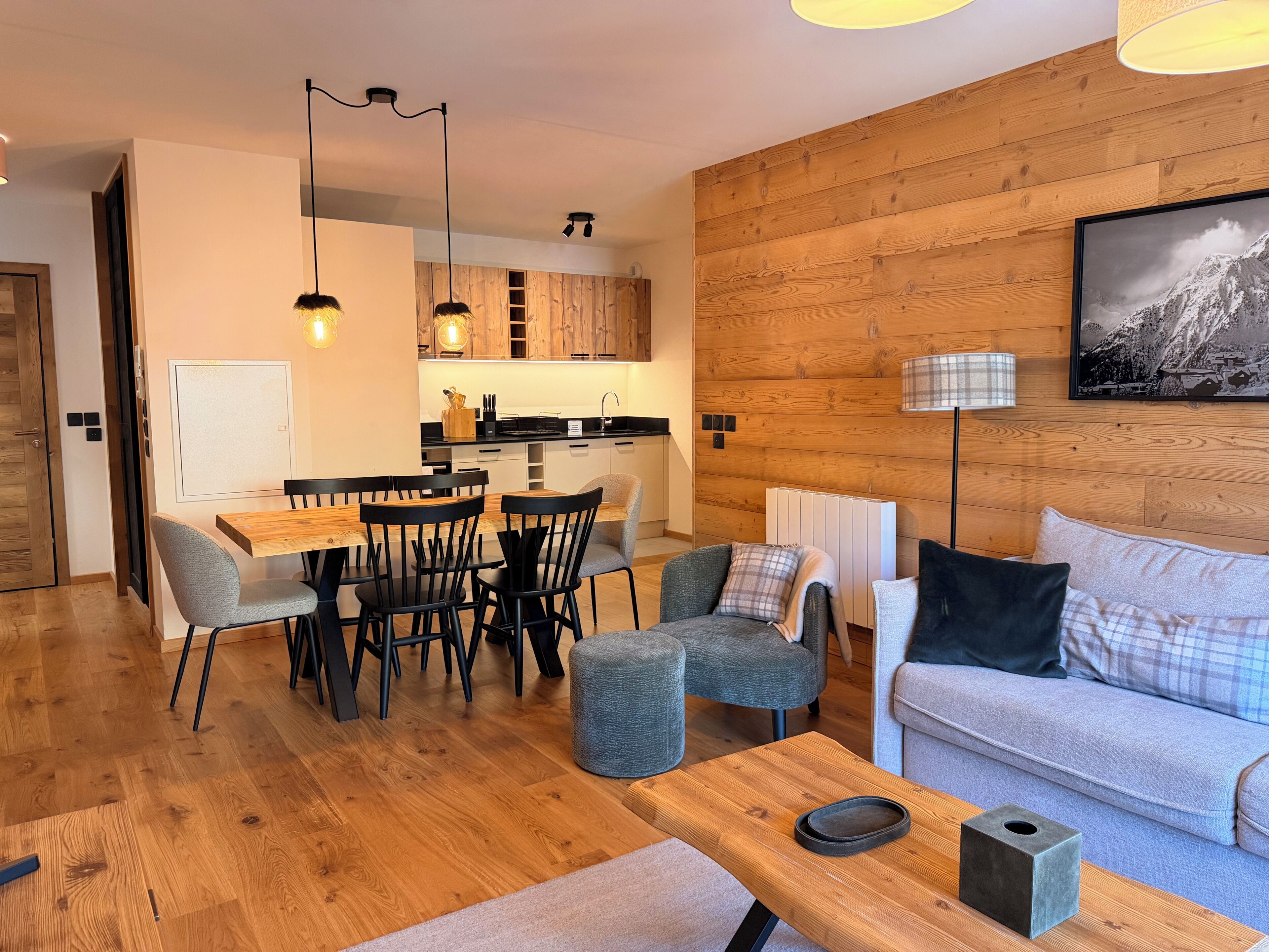 A modern living room with a wooden floor, a grey sofa, a dining table with chairs, and a kitchen in the background.