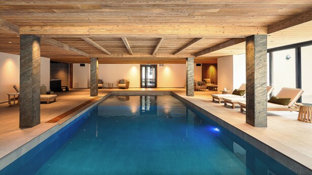 A swimming pool with wooden beams and stone pillars, surrounded by lounge chairs and a sunlit room. - Résidence l'Alpeggio by My Mountain Resort (Les Deux Alpes)