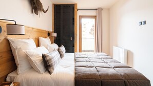 A bedroom with a wooden headboard, a bed with white and plaid bedding, a lamp, a wall-mounted heater, and a door leading to a balcony. - Résidence l'Alpeggio by My Mountain Resort (Les Deux Alpes)