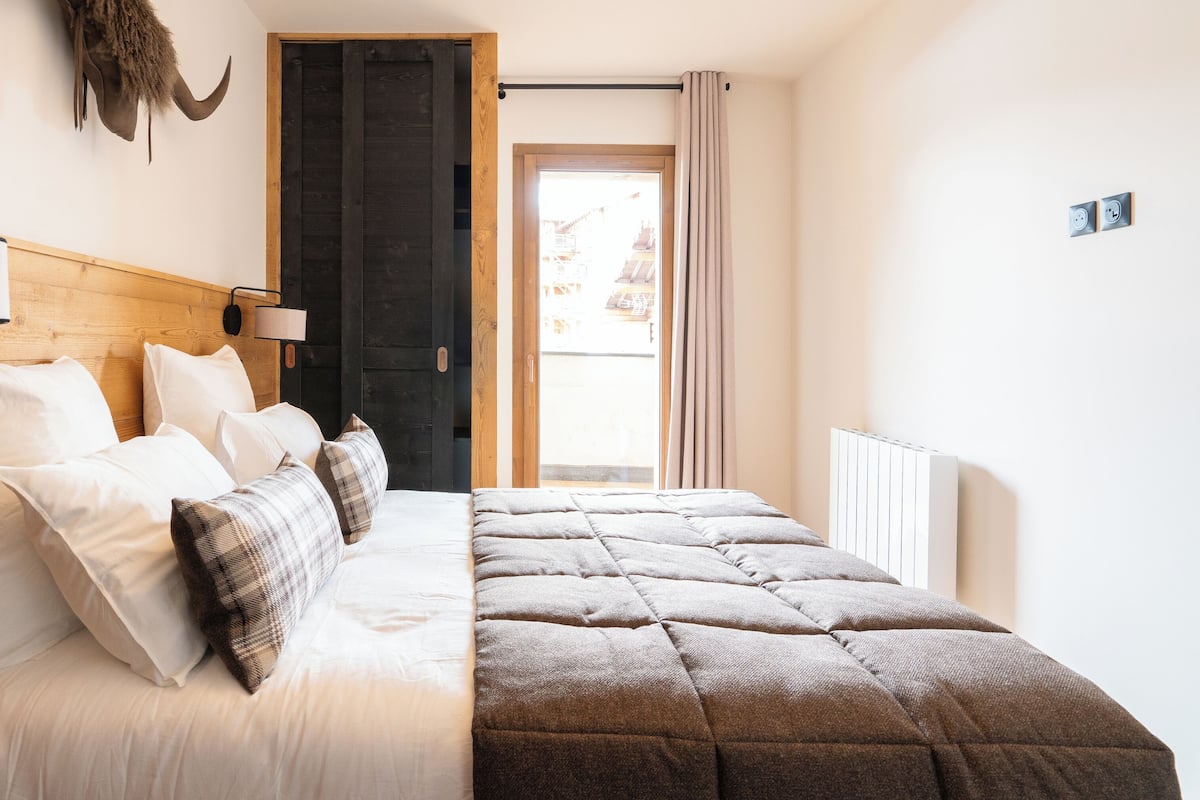 A bedroom with a wooden headboard, a bed with white and plaid bedding, a lamp, a wall-mounted heater, and a door leading to a balcony.