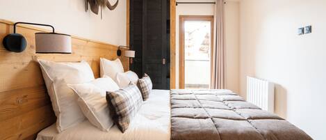 Un dormitorio con cabecera de madera, cama con ropa de cama blanca y de cuadros, una lámpara, un calentador de pared y una puerta que da a un balcón.