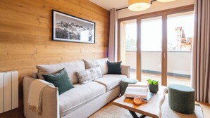 A modern living room with a wooden wall, a sofa with cushions, a coffee table with books and a plant, and a large window with curtains. - Résidence l'Alpeggio by My Mountain Resort (Les Deux Alpes)