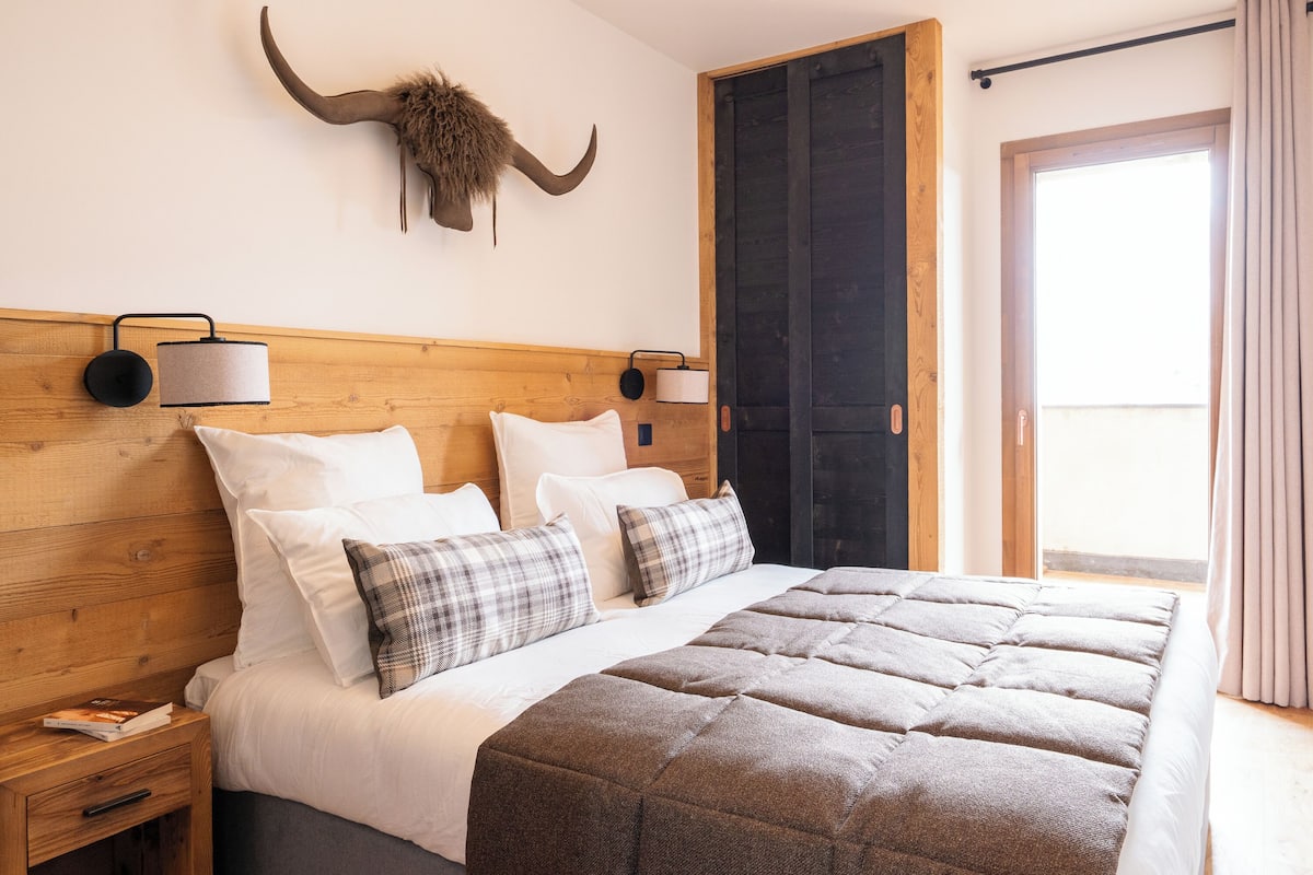 A bedroom with a wooden headboard, a large bed with a brown quilt, two bedside lamps, a nightstand, a wall-mounted light fixture, and a door with a curtain.