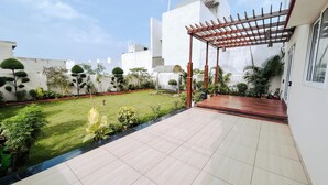 A modern patio with a wooden pergola, a well-maintained lawn, and a variety of plants. - Shyamsudha Homestay (Ujjain)