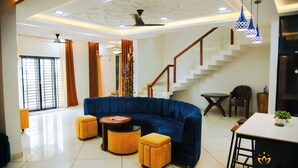 A spacious living area with a blue sectional sofa, a round coffee table, and a dining table with bar stools. A staircase with a wooden railing leads to an upper floor. - Shyamsudha Homestay (Ujjain)