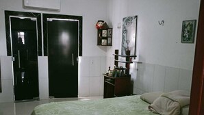 Un dormitori amb un llit, una tauleta de nit, una estantanya penjada a la paret amb objectes decoratius i una porta.